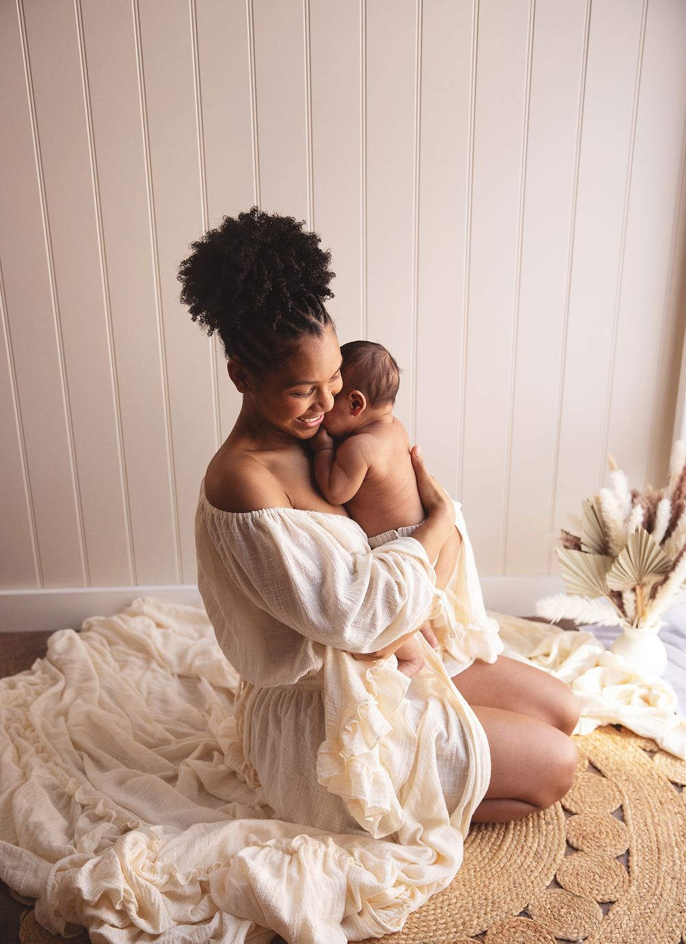 Mother in white dress holding newborn on woven rug with dried pampas grass decor and beige panelled wall. During a newborn shoot in Bexley