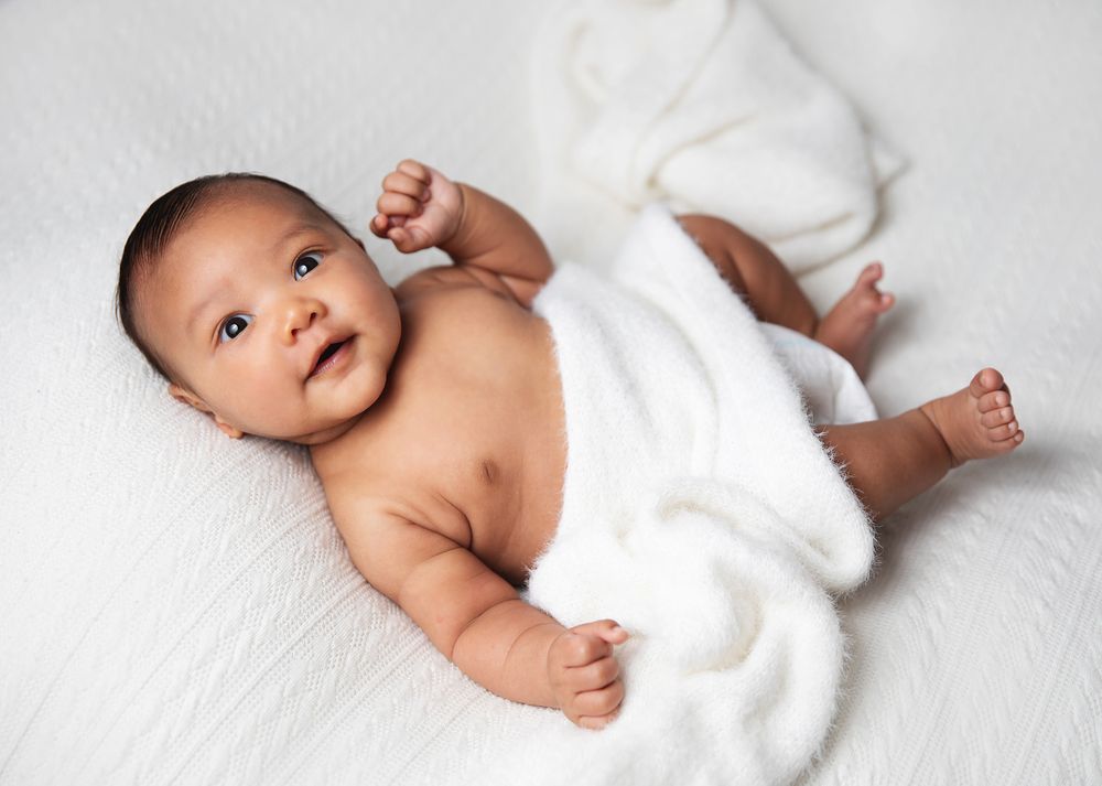 Baby wearing white wrap lies on white bedding while looking toward camera with arms outstretched. During a newborn photoshoot in London