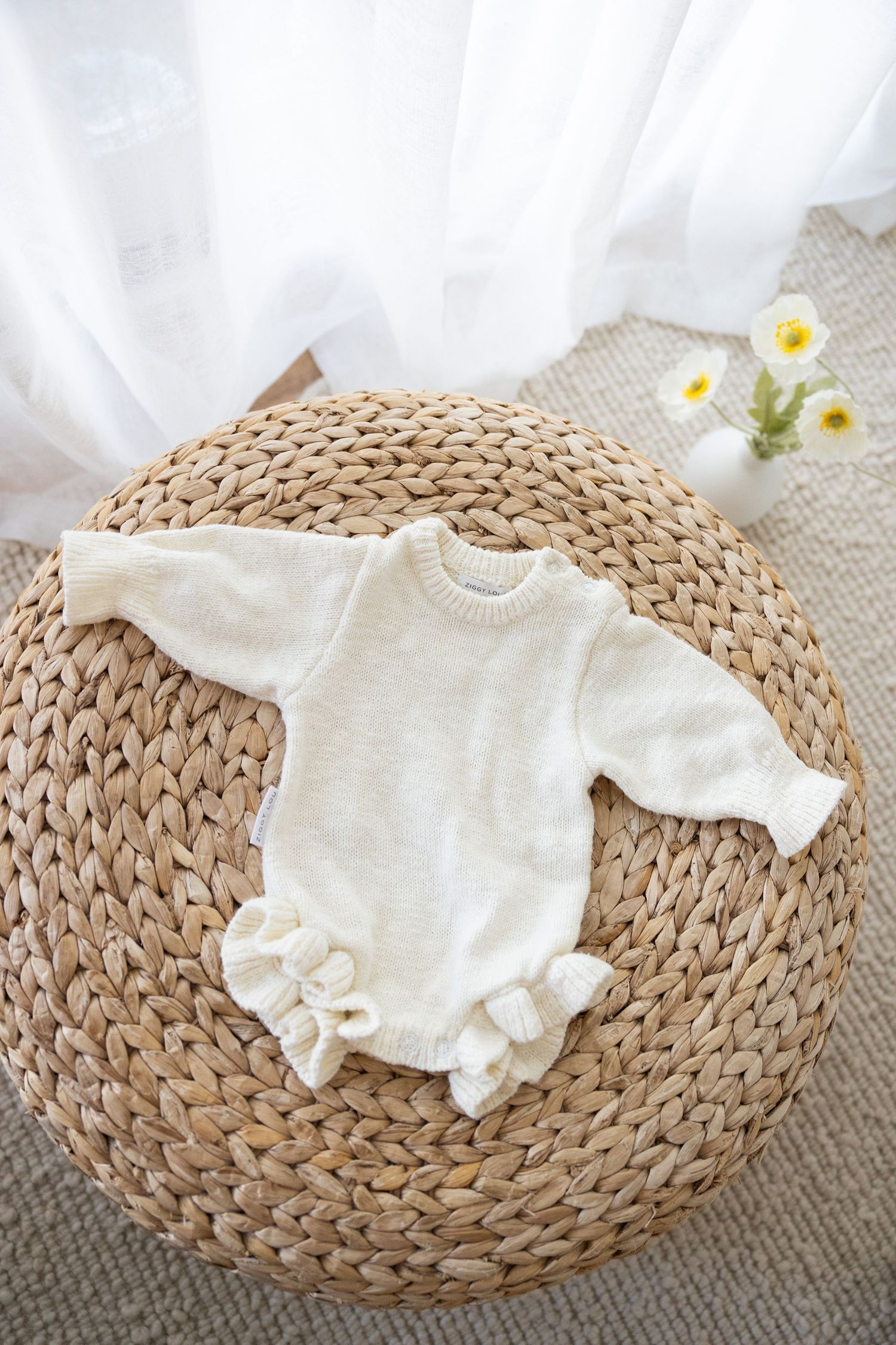 A white knitted baby romper with ruffled trim laid on a round woven straw mat with small daisies in the background.