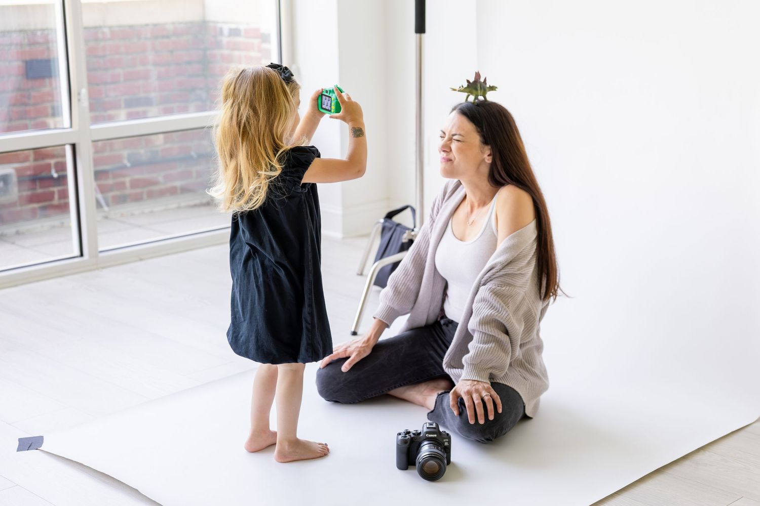 A small child in a dress holds up a toy camera while someone sits cross-legged on a bright white floor by windows with a toy dinosaur.