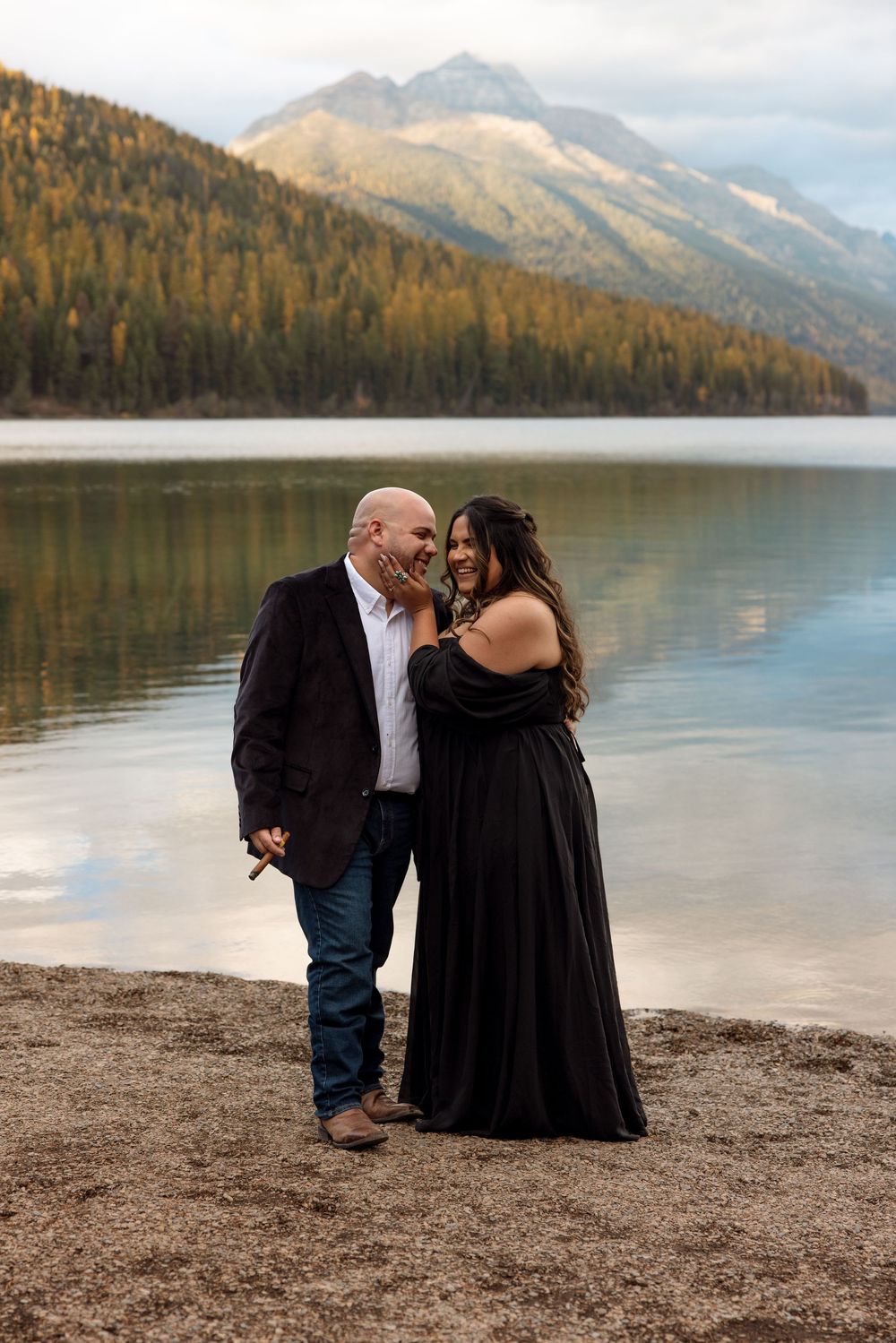A couple embraces by a serene mountain lake with autumn forests and peaks in the background.