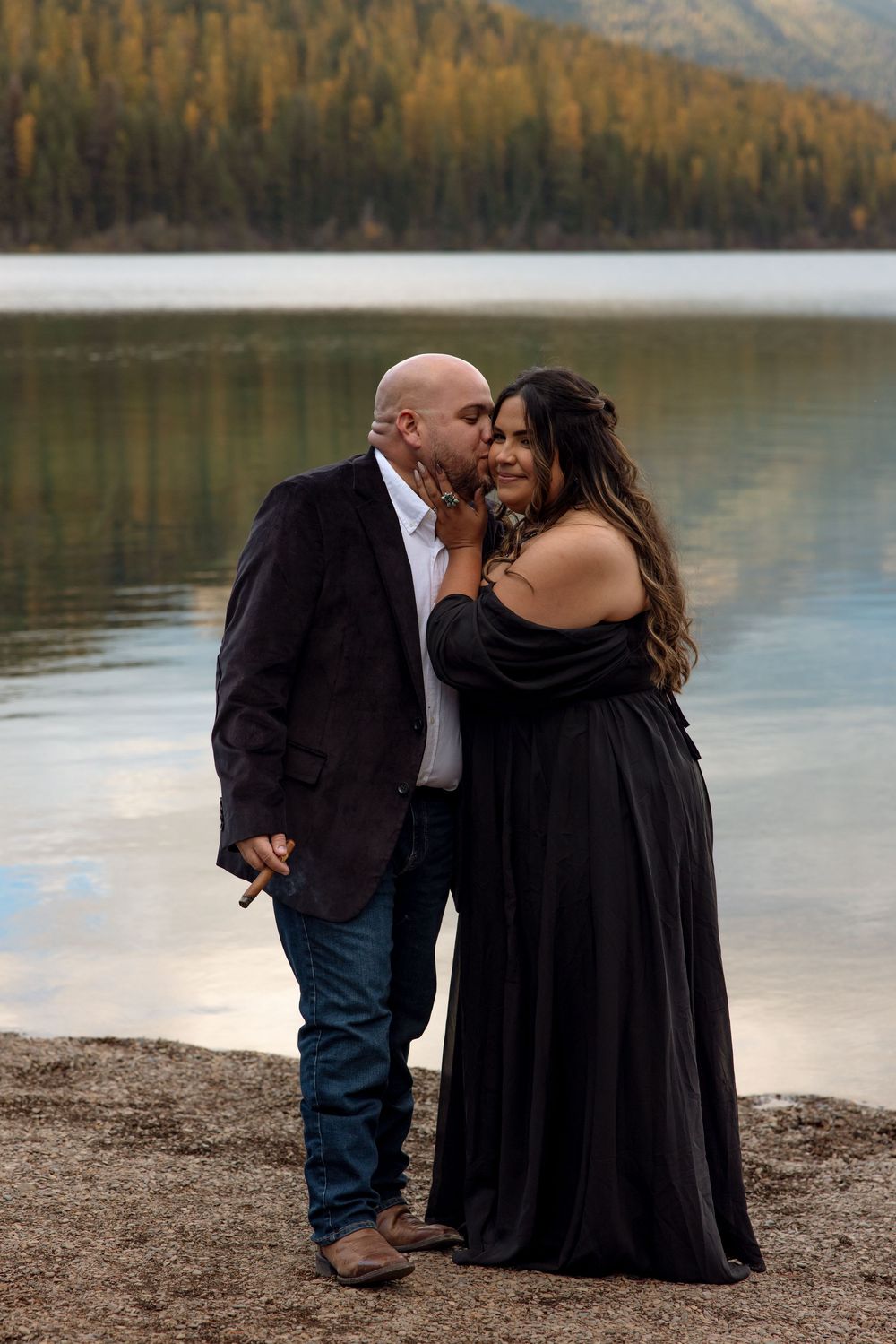 Romantic couple in black formal attire sharing an intimate moment by a serene mountain lake at sunset.