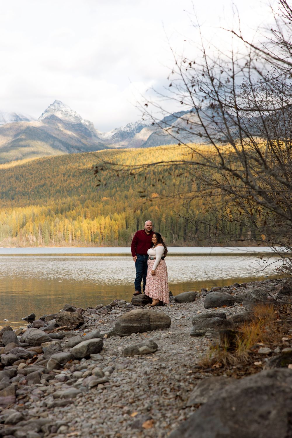 A couple stands on rocky lakeshore with snow-capped mountains and golden autumn forest in background at dusk.