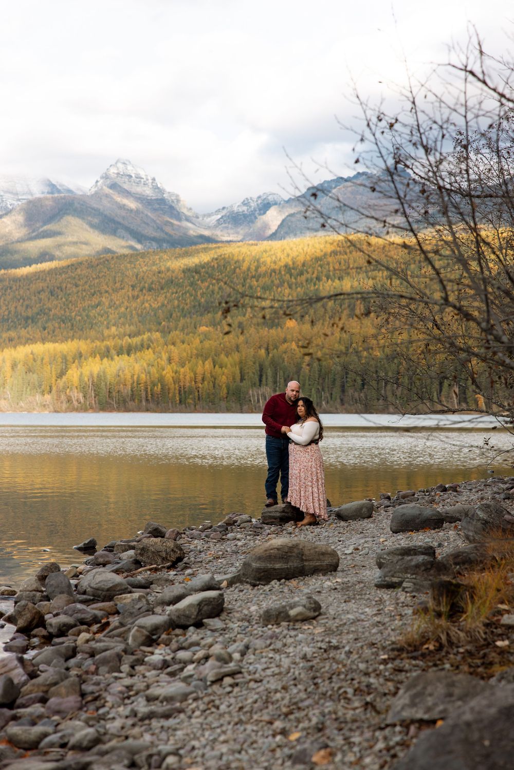 Two figures stand on rocky shoreline of mountain lake with autumn forest and snow-capped peaks in background.