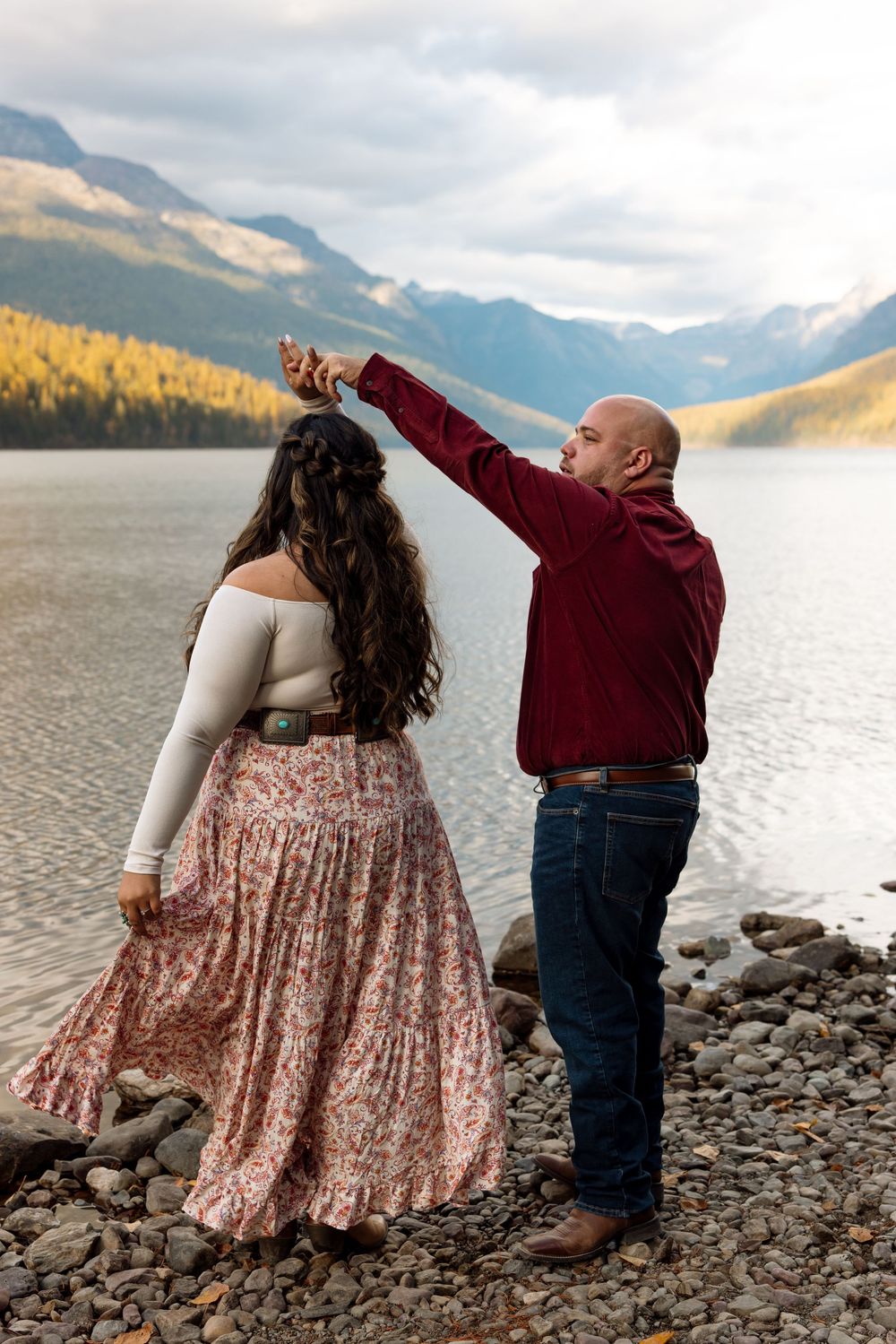 A couple dances together on a rocky lakeshore with mountains and autumn trees in the background.