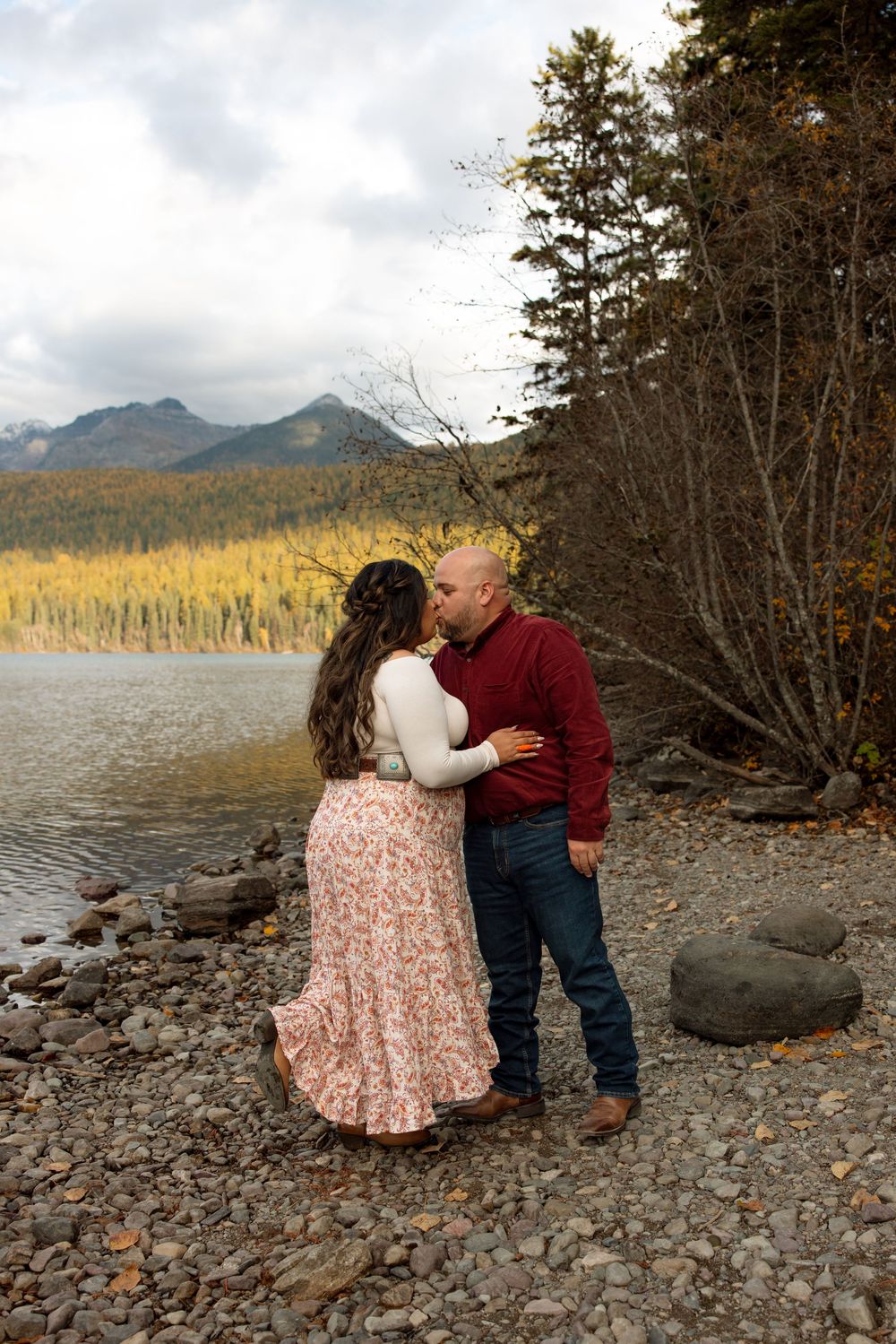 A couple shares a romantic moment by a mountain lake with autumn trees and rocky shoreline in the background.