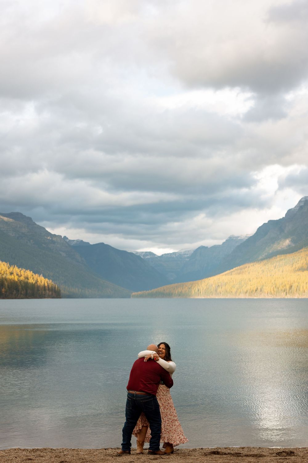 Couple embraces while standing on shore of mountain lake with dramatic clouds and golden sunlit forests in background.