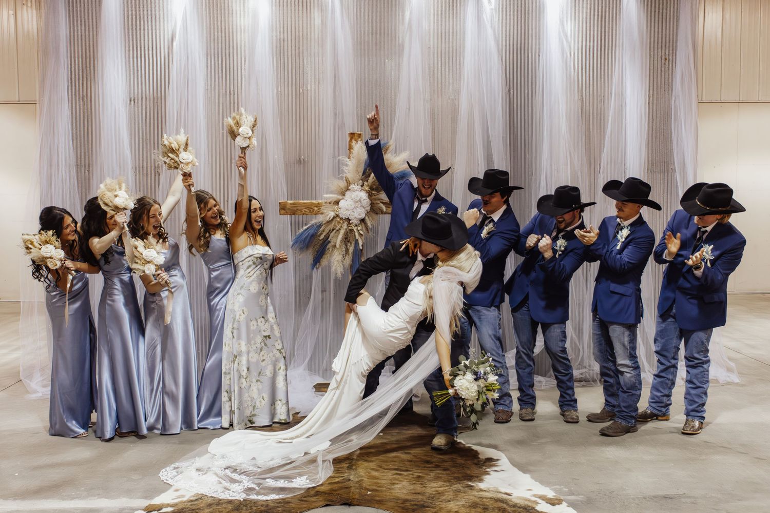 Wedding party in blue dresses and cowboy attire poses dramatically against white draped backdrop during celebration.