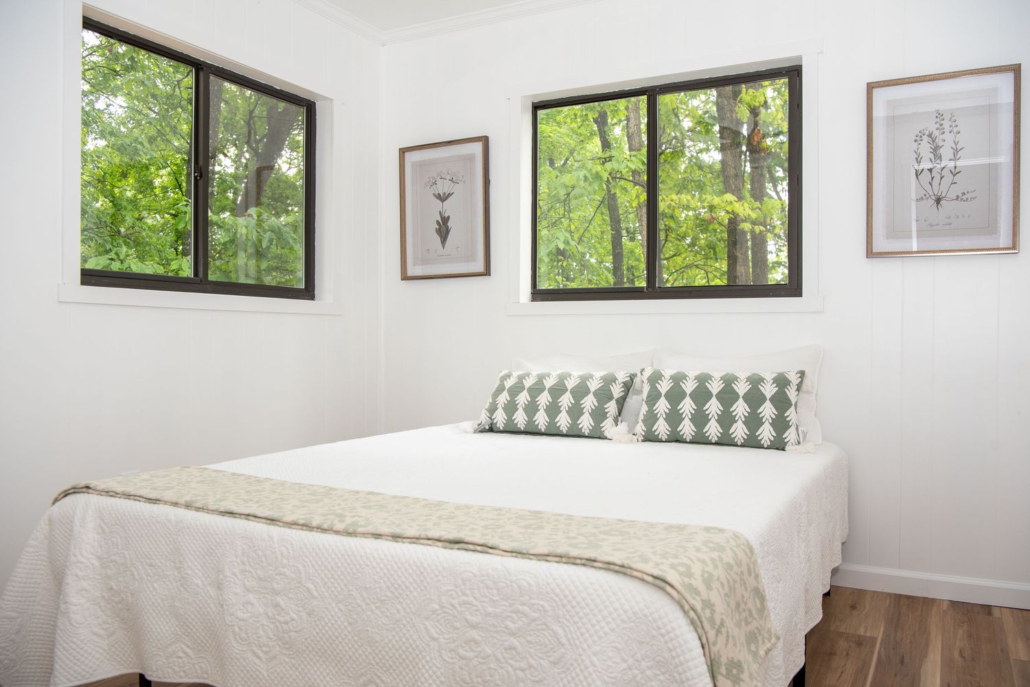 Bright modern bedroom with white bedding and walls features multiple windows showcasing forest views.