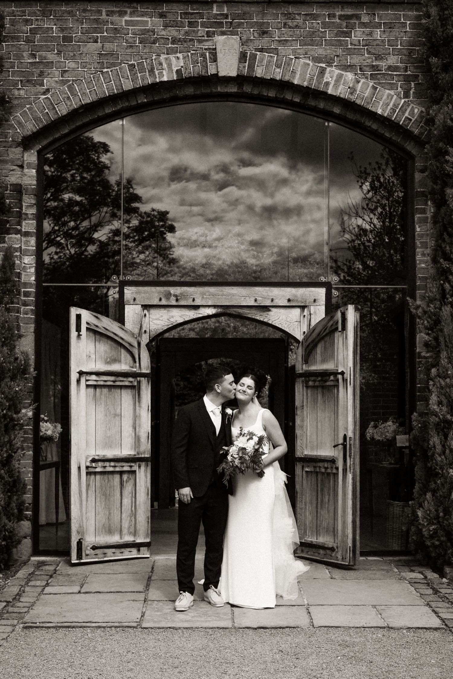 A couple stands in an arched brick doorway with rustic wooden doors opened wide on their wedding day.