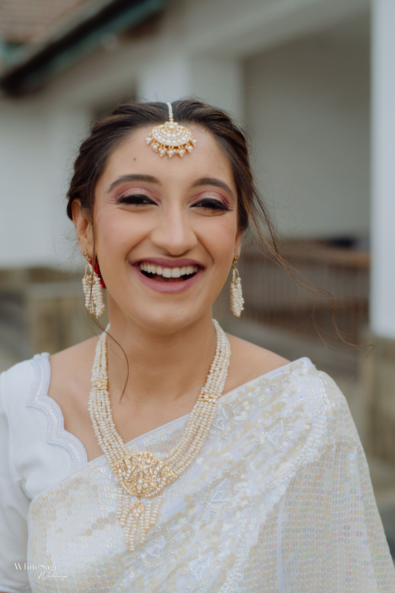 The bride wears elegant pearl jewelry and a white sari with traditional Indian maang tikka headpiece while smiling warmly.