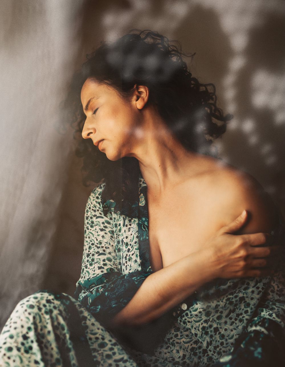 Person wearing a floral patterned dress poses artistically in dim lighting against a blurred background.