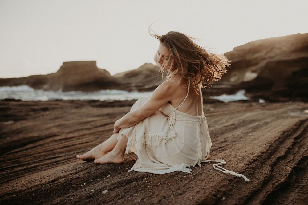 Woman sitting on dark rocky ground in flowing dress at sunrise.