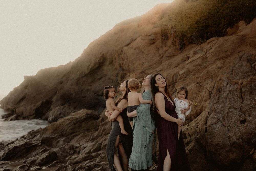 Women holding toddlers against dramatic coastal cliff at golden hour.