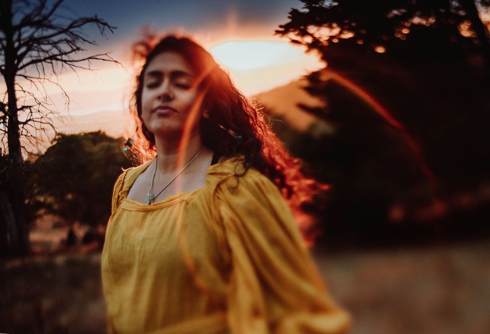 Woman in yellow dress backlit by vibrant sunset through trees.