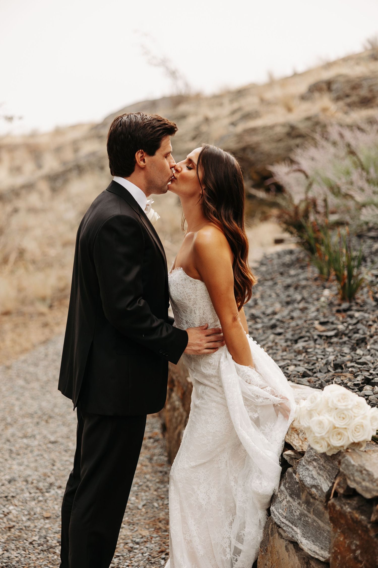 A bride in a flowing mermaid wedding dress stands on a wooden deck overlooking mountains and water as her veil catches the wind.