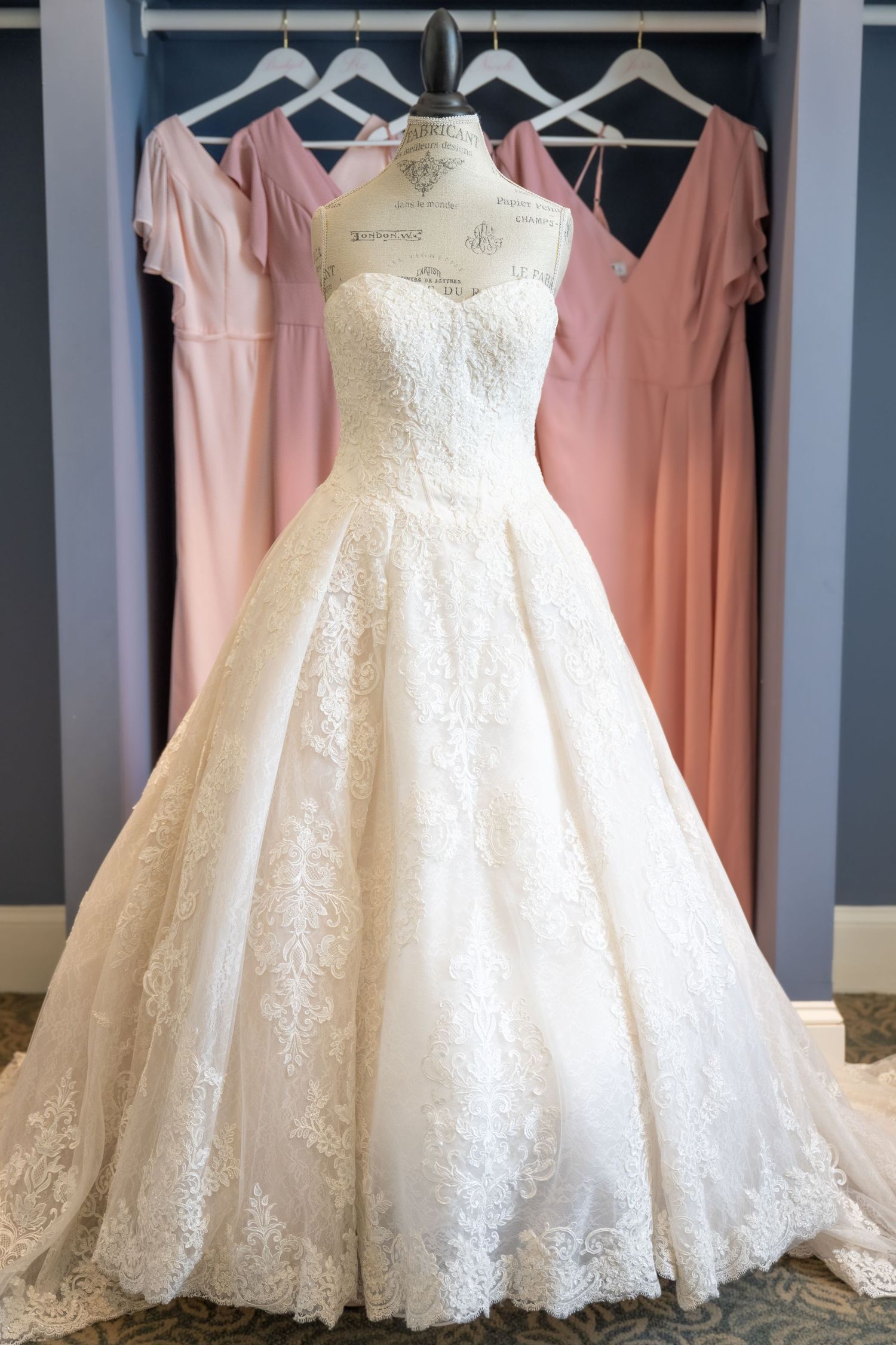 Elegant strapless lace ball gown wedding dress on mannequin with pink bridesmaid dresses hanging in background.