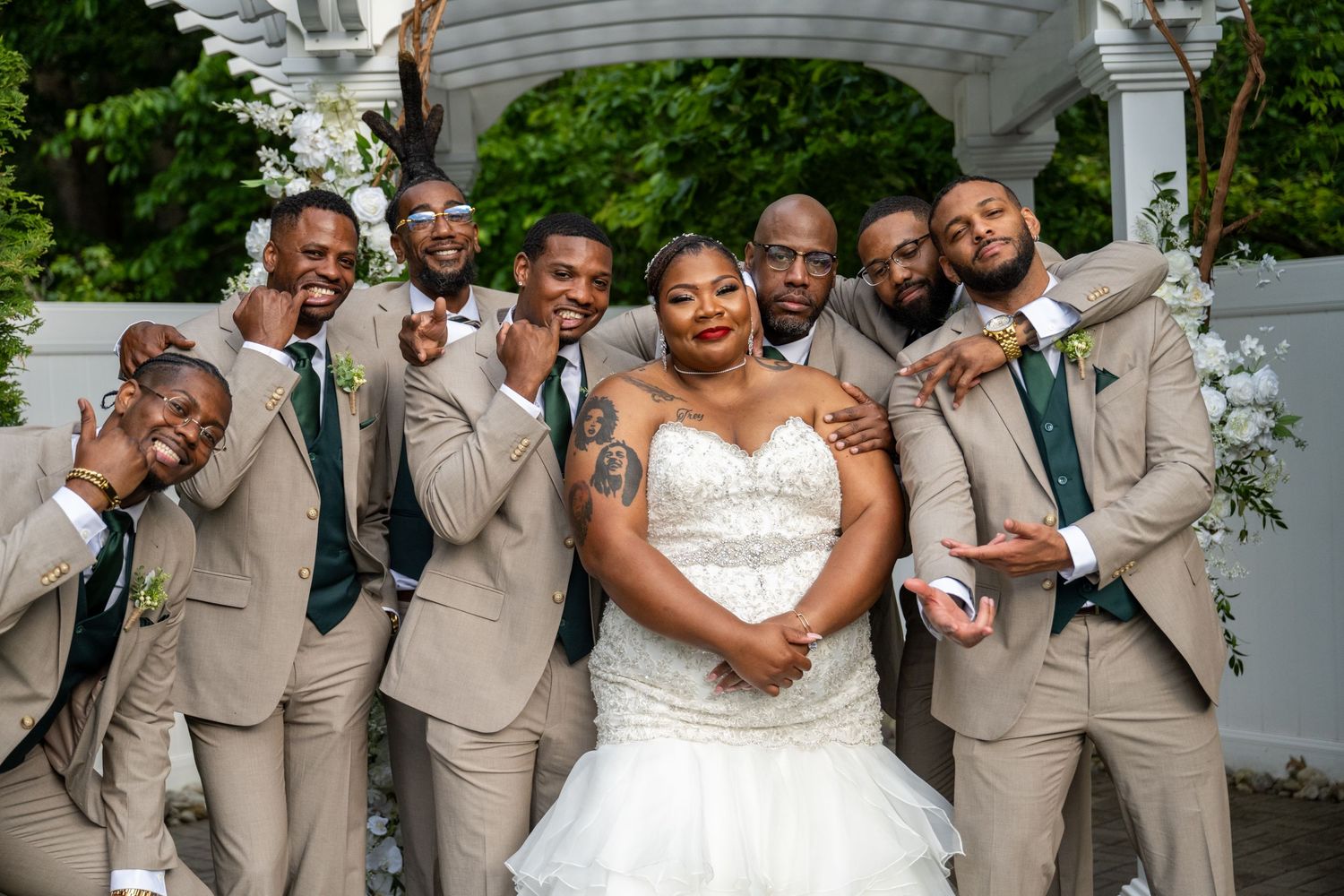 Bride in white gown poses with groomsmen in tan suits and green ties at outdoor wedding ceremony.