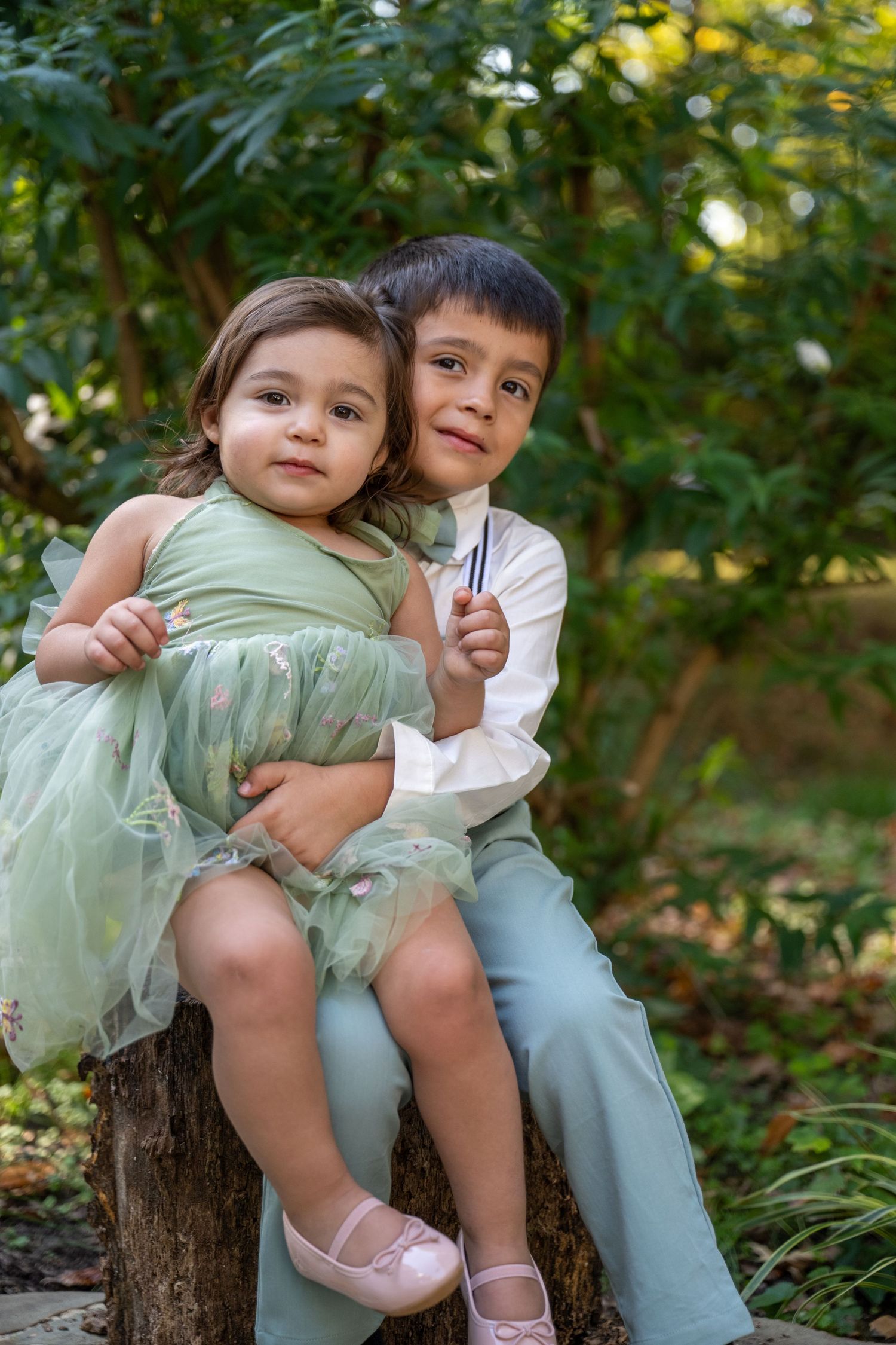Two young children in formal attire sit together on a wooden stump in a garden setting.