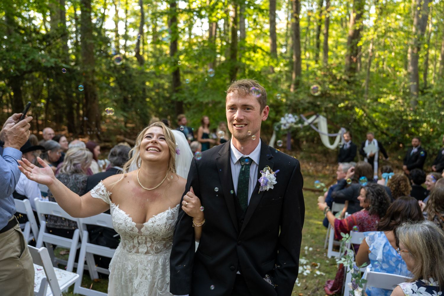 Two newlyweds walk down the aisle while guests seated on white chairs celebrate during an outdoor forest wedding ceremony.