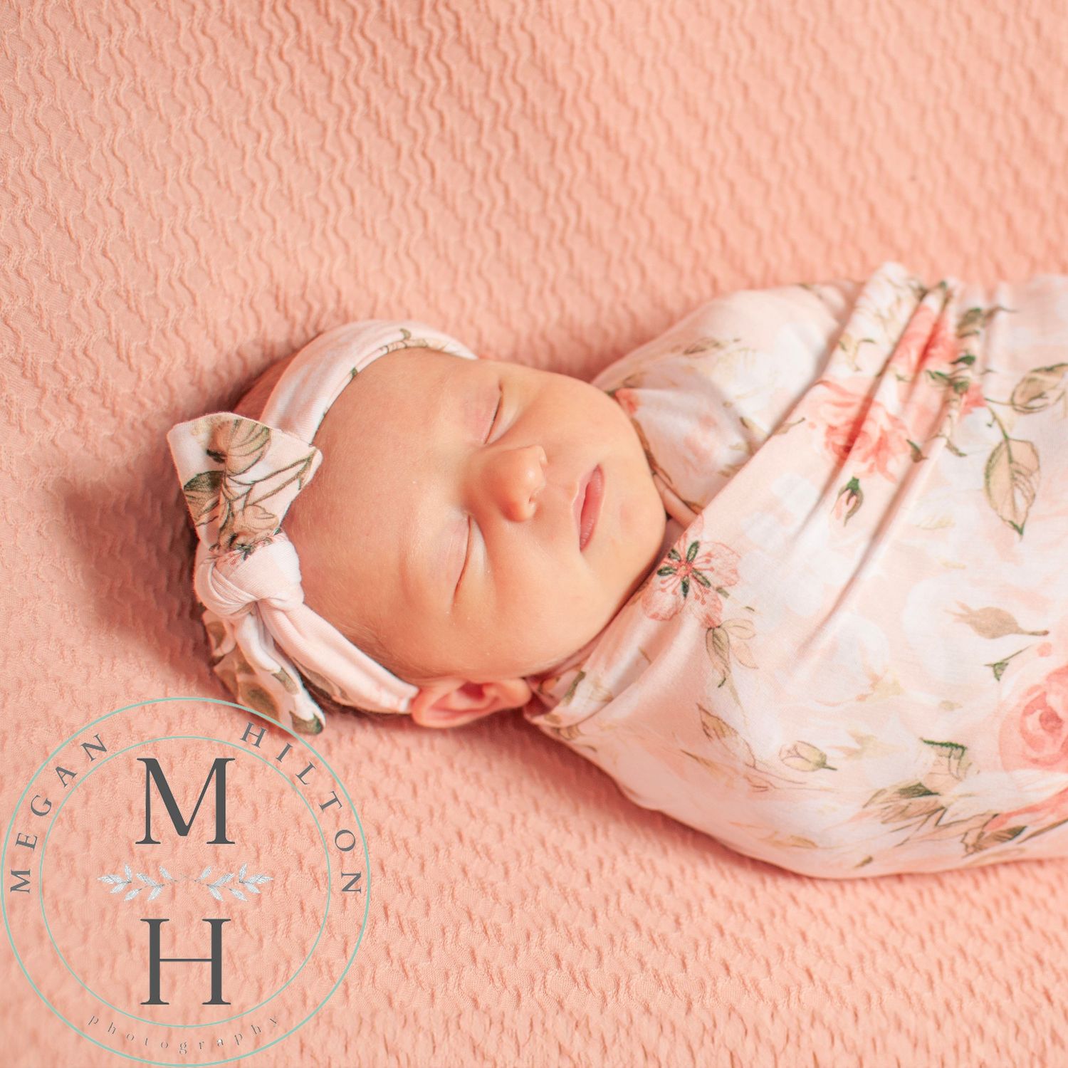 Peacefully sleeping newborn swaddled in floral print wrap on peach textured background.