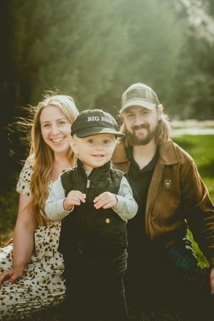 Family portrait outdoors in natural lighting with child wearing a baseball cap standing between parents.