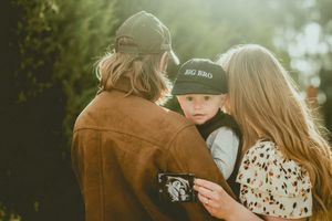 A toddler in a baseball cap being held outdoors in sunlit natural setting.