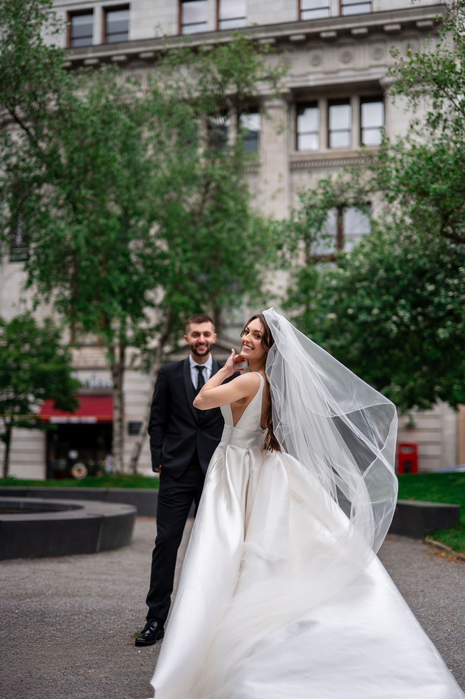 Bride in flowing white gown and veil smiles as groom in black suit walks behind her outdoors near green trees.