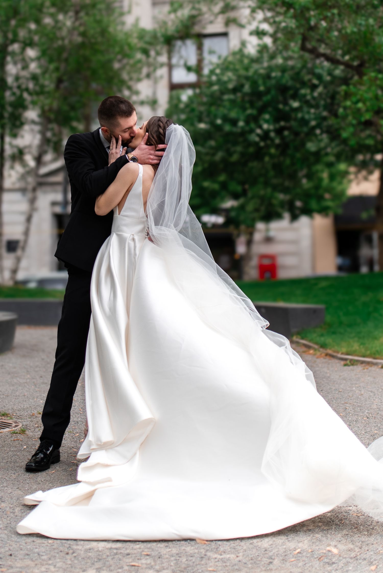 A bride and groom share a romantic kiss outdoors, her white gown flowing gracefully as he holds her face tenderly.