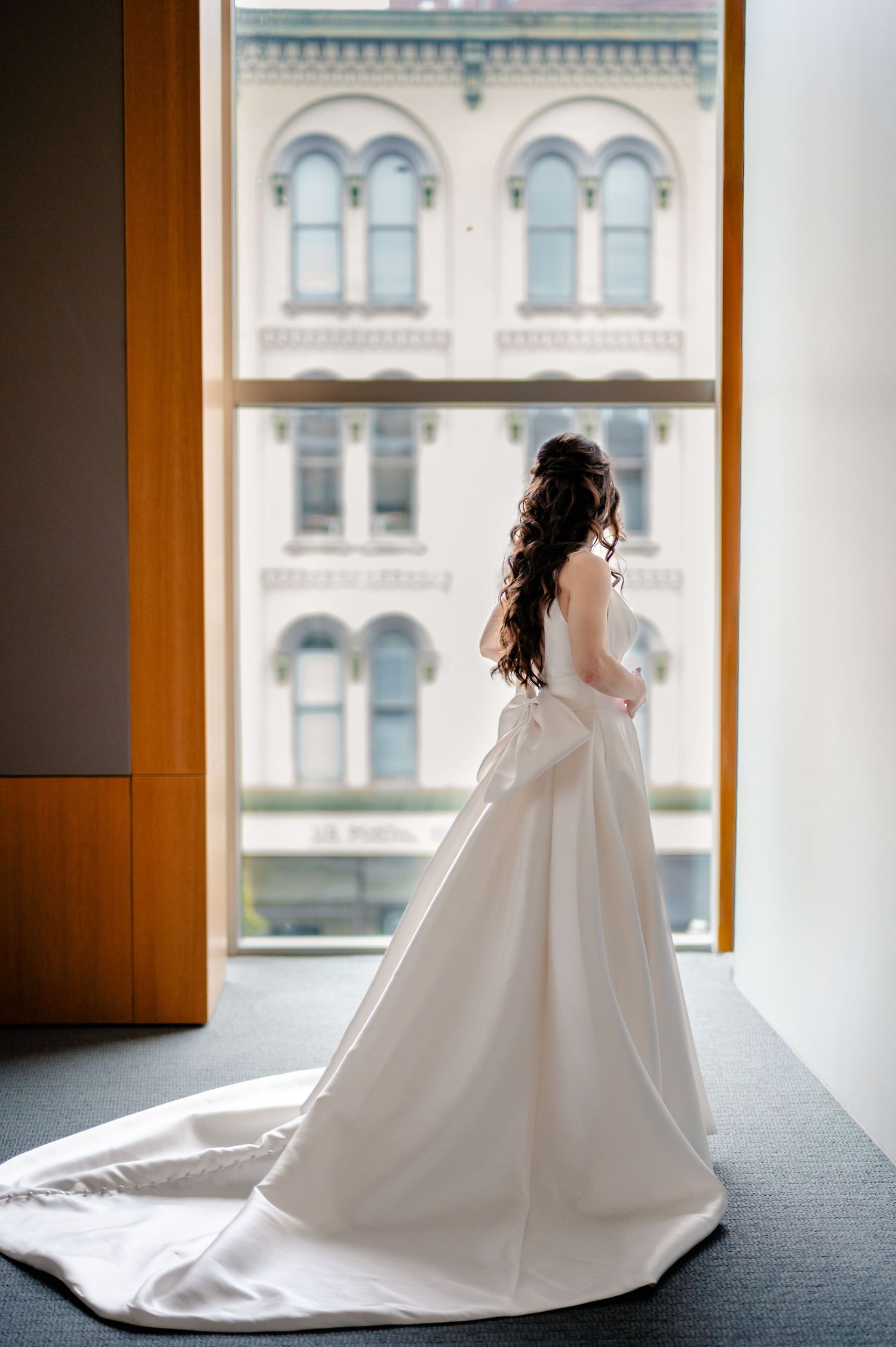 Bride in elegant white ball gown with long train stands by large window overlooking historic building.
