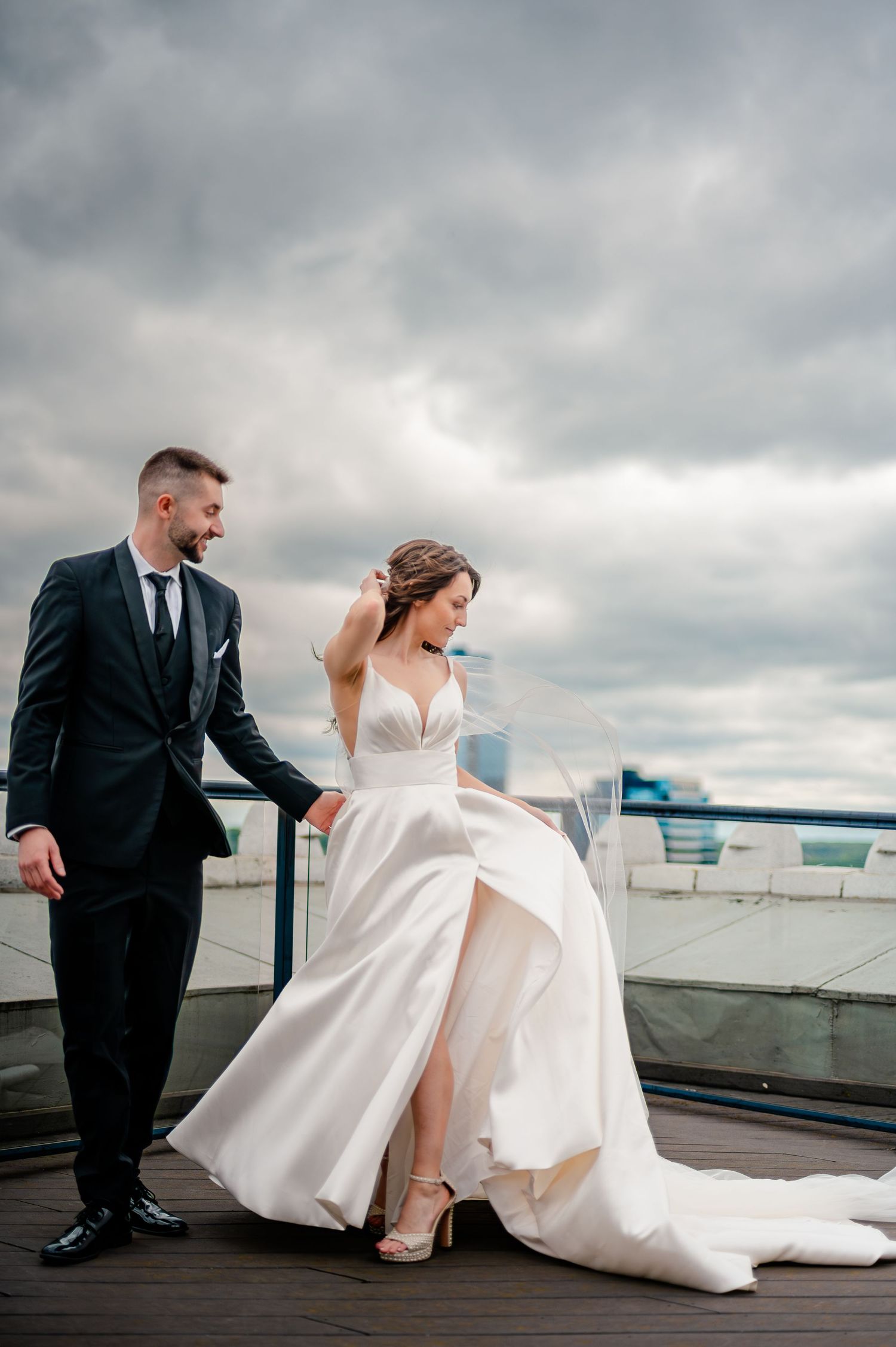 Bride in white gown and groom in black suit pose on rooftop terrace under dramatic cloudy sky.