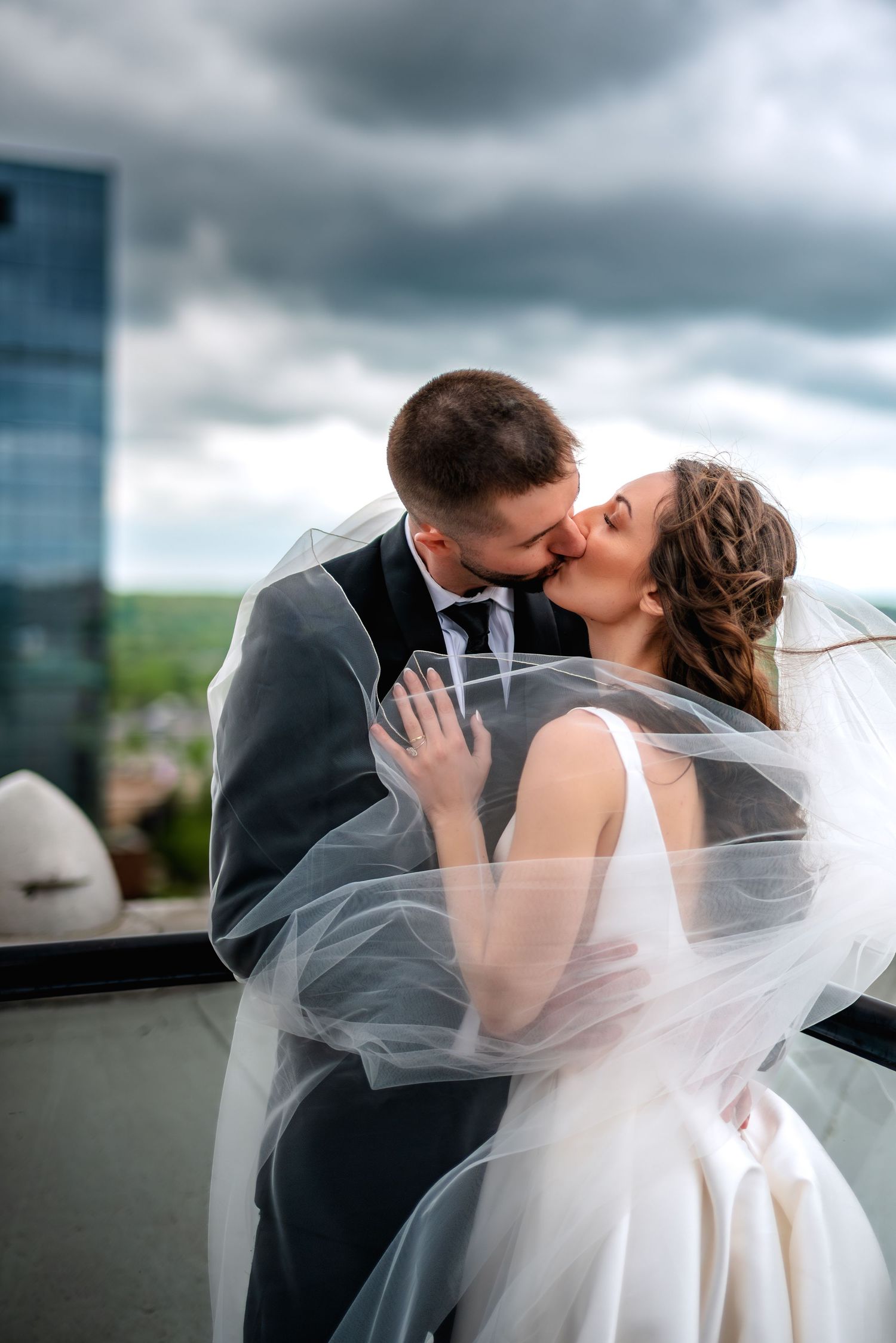 A bride and groom share a romantic kiss outdoors on a rooftop, her veil flowing in the wind against a cloudy sky.