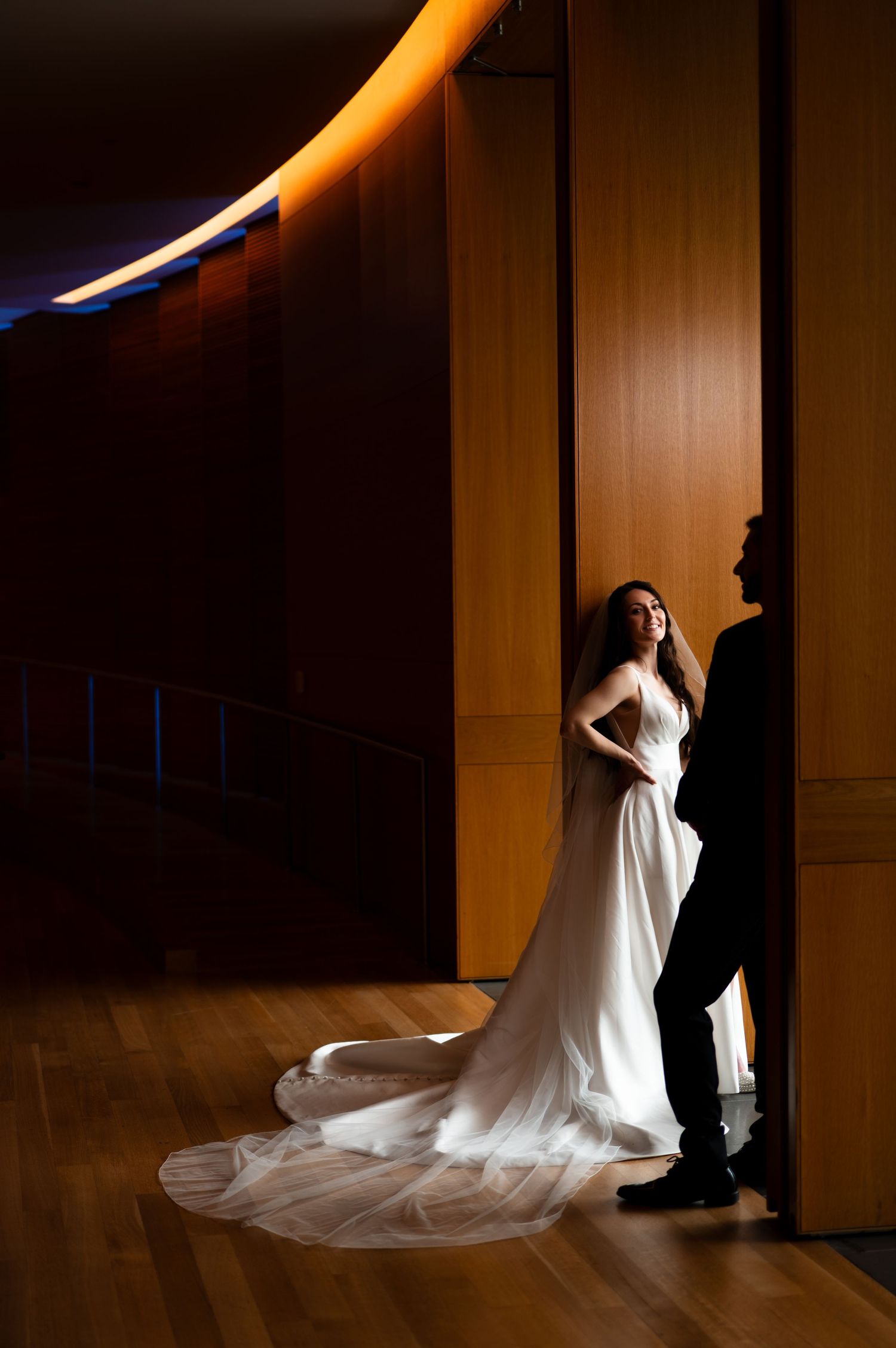 Bride in elegant white gown with long train stands in dramatic light beside groom in modern wood-paneled venue.
