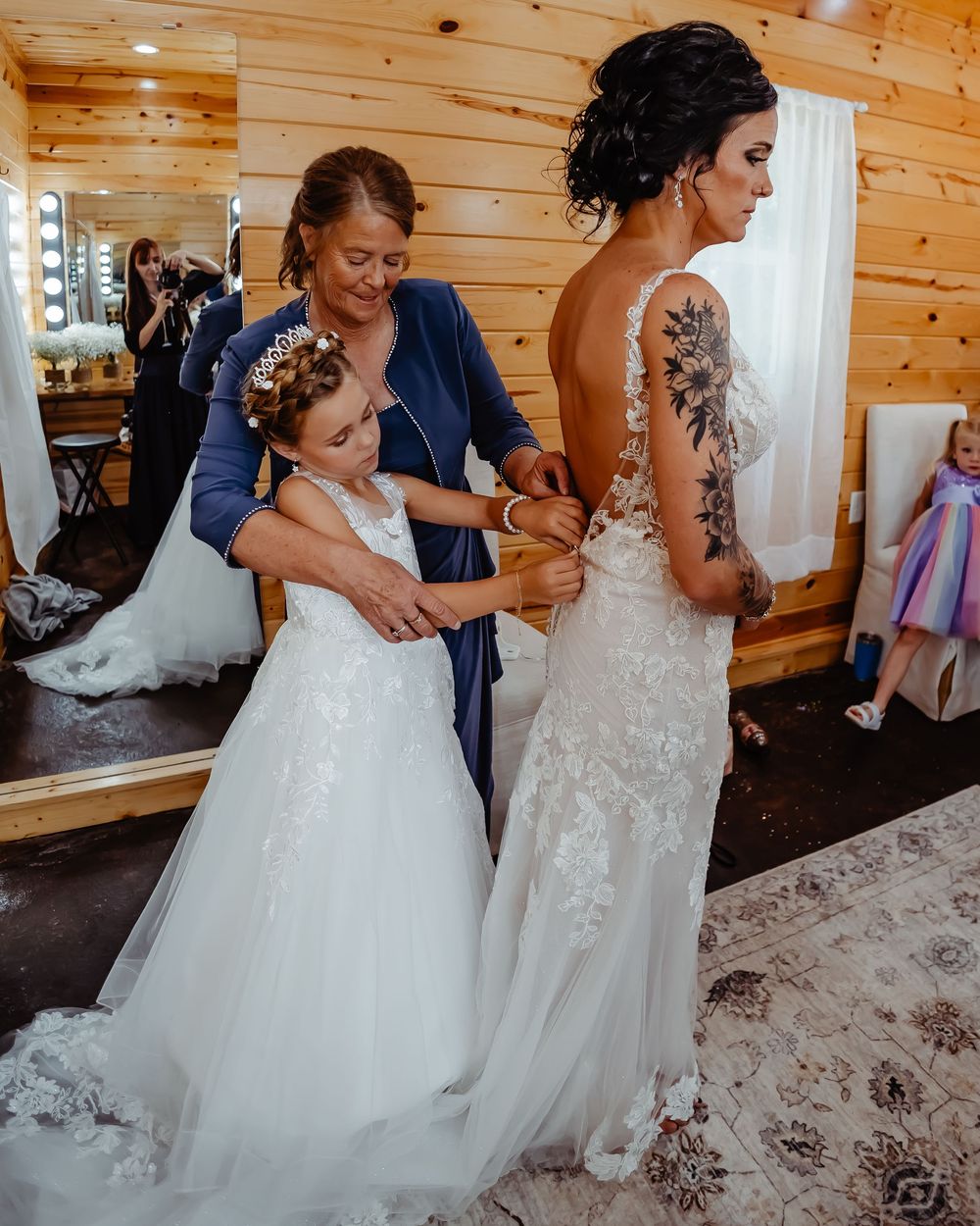 Two elegant wedding dresses being adjusted in a rustic wood-paneled bridal suite with a large mirror.