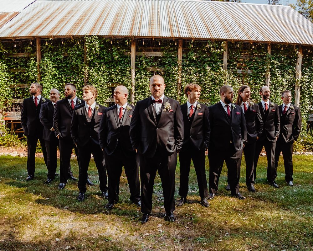 Groomsmen in black suits pose for formal photo under metal gazebo covered in greenery.
