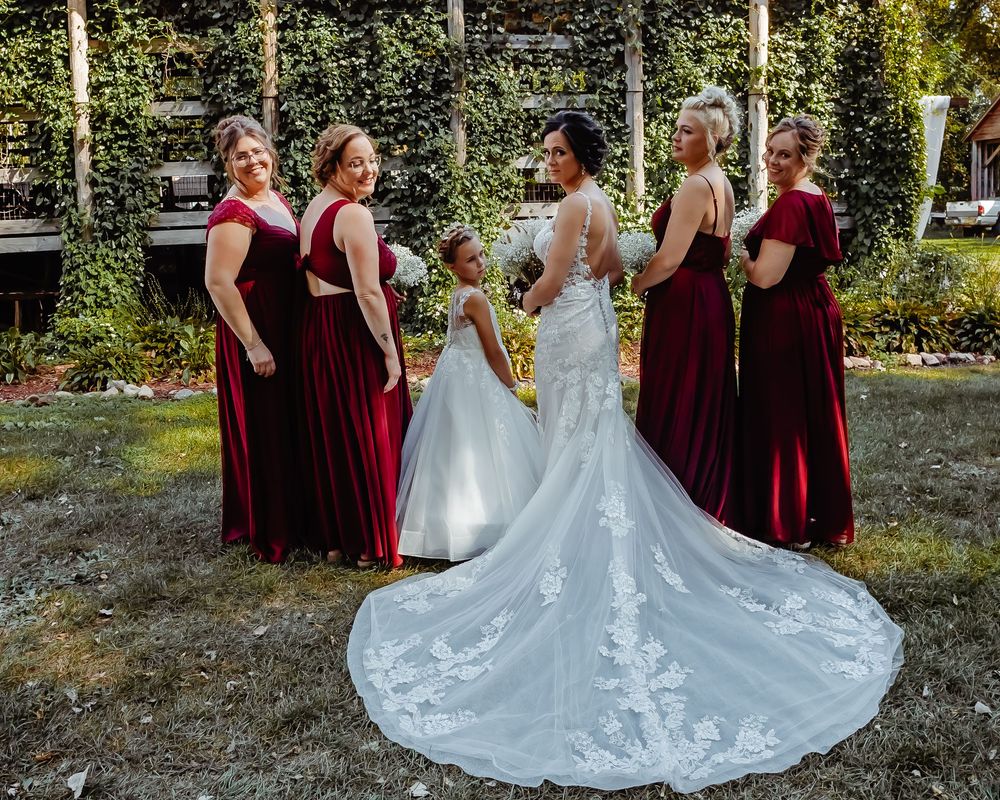 Wedding party in burgundy dresses and white gown posed together outdoors against natural greenery.
