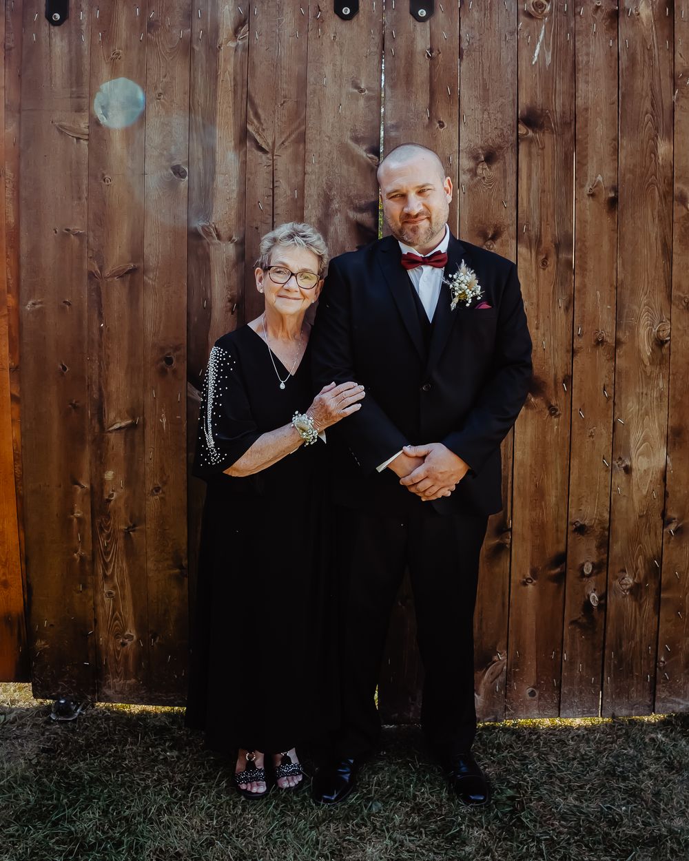 Wedding guests in formal attire standing against rustic wooden fence backdrop.