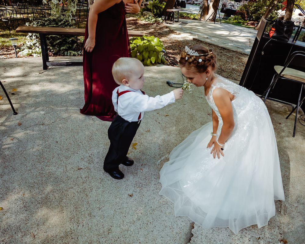 A small child in formal wear dances with a person in a white wedding dress on a paved garden path.