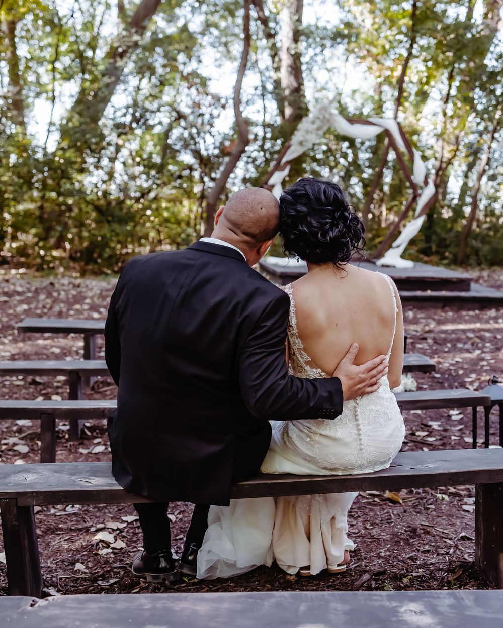 A couple in wedding attire share an intimate moment while sitting on a wooden bench in a wooded setting.