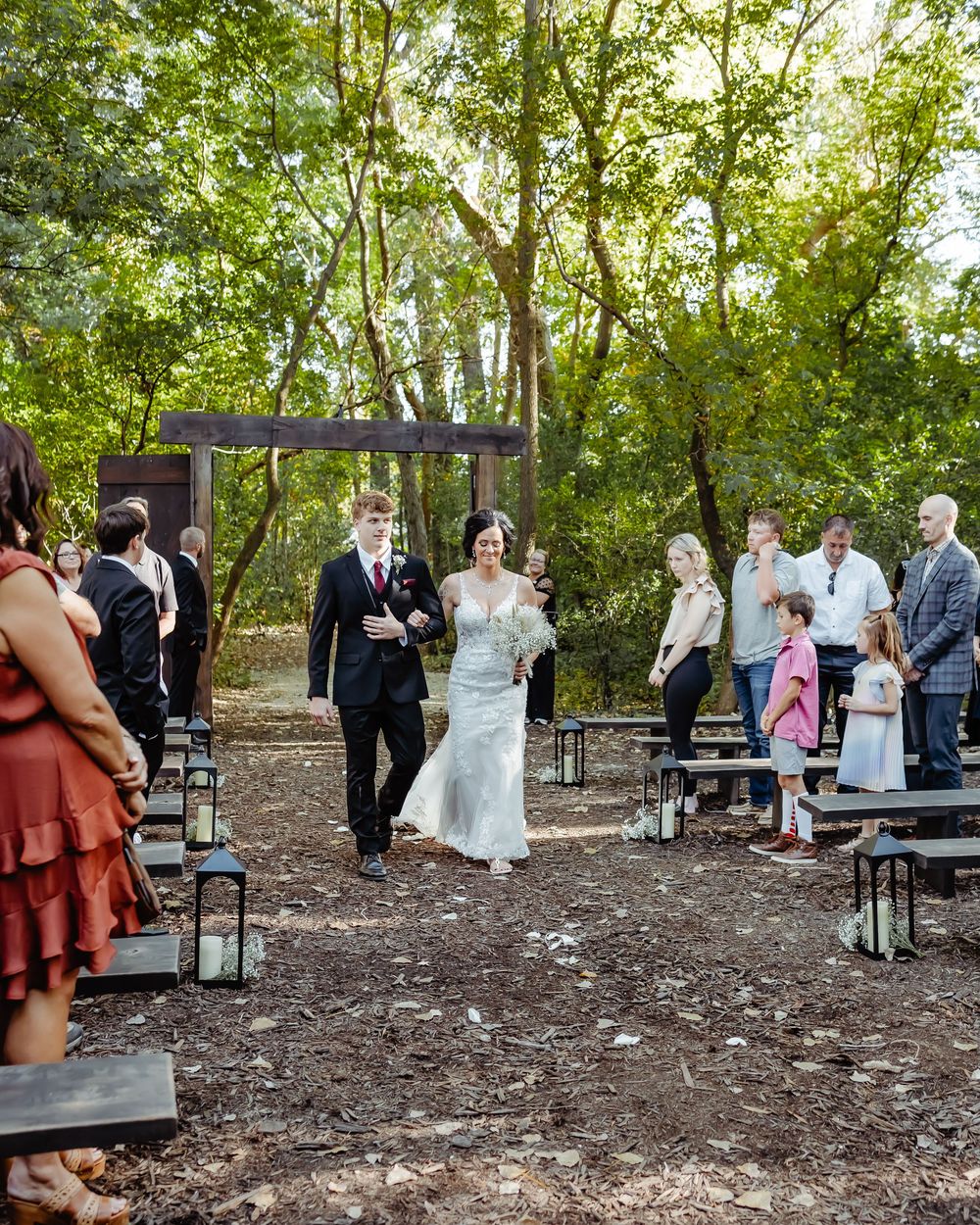 Outdoor woodland wedding ceremony with rustic wooden benches and lanterns lining the aisle.