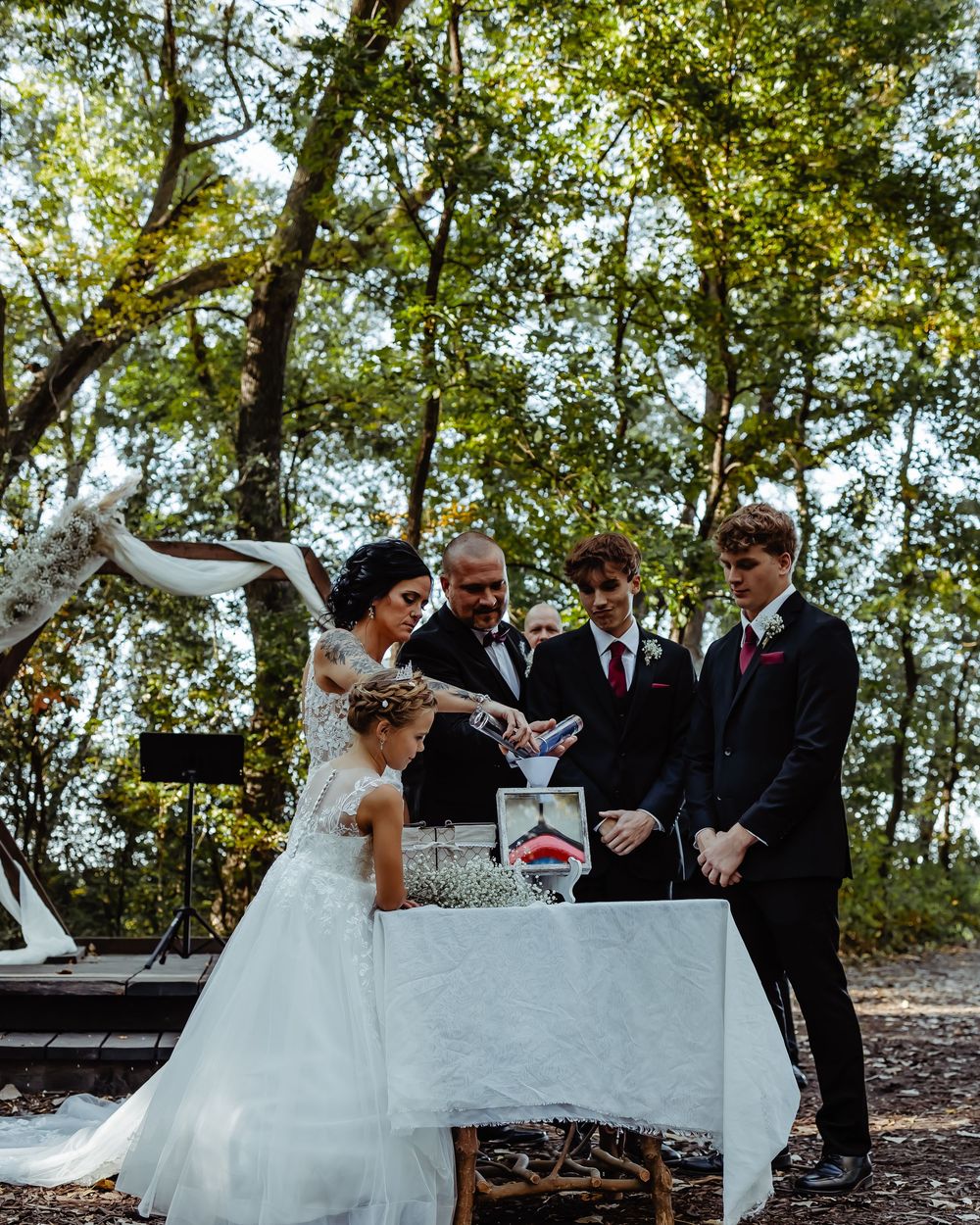 Wedding ceremony shows couple performing unity ceremony at outdoor woodland altar.