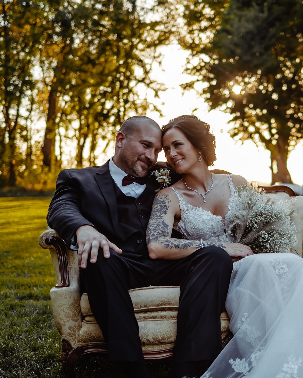 A couple in wedding attire share a tender moment on a vintage chair at sunset.
