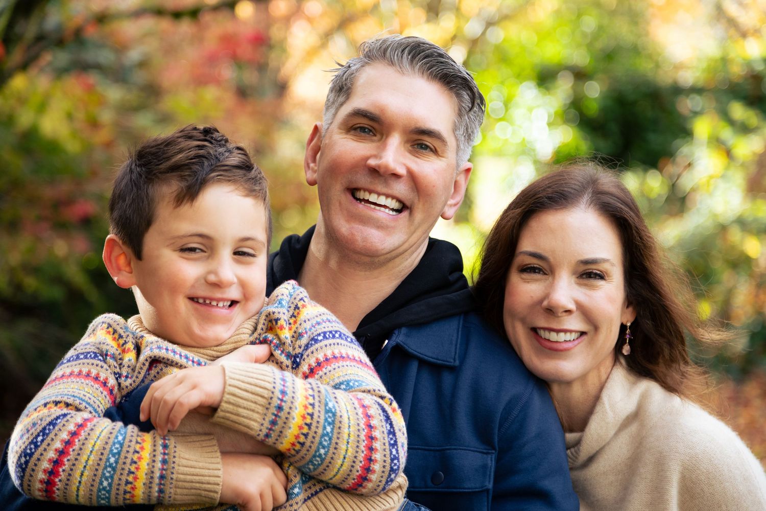 Happy family of three smiling outdoors in autumn, parents and young boy in colorful sweater posing together.