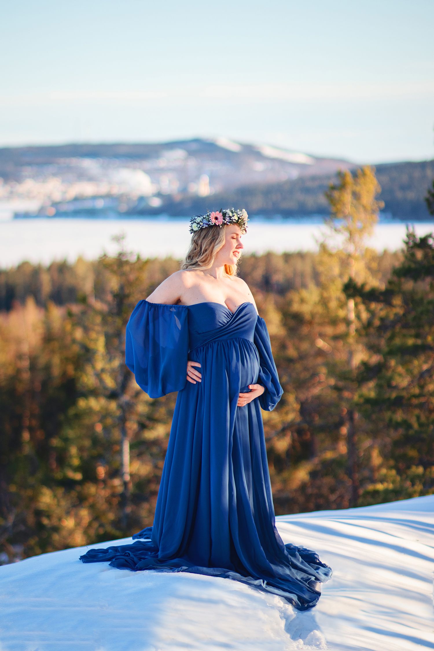 Pregnant woman in blue flowing gown with floral crown standing on snowy hilltop with forest view.