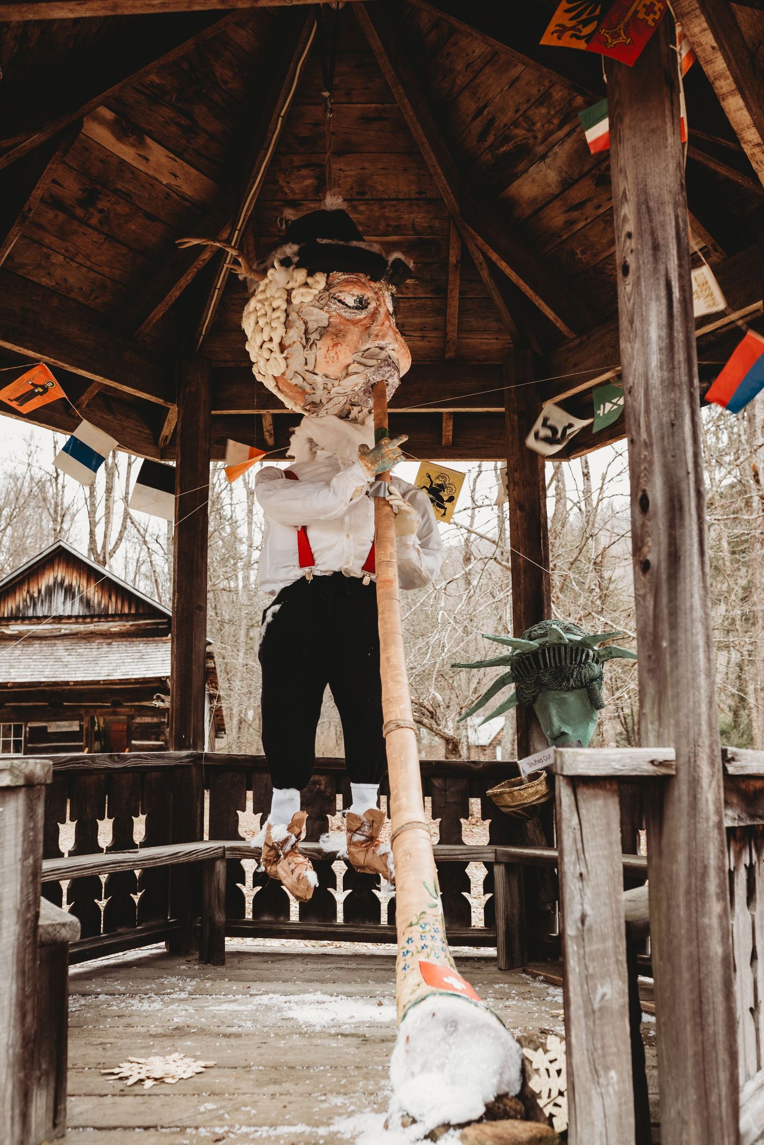 Whimsical scarecrow figure with vintage styling displayed in rustic wooden gazebo with winter forest backdrop.