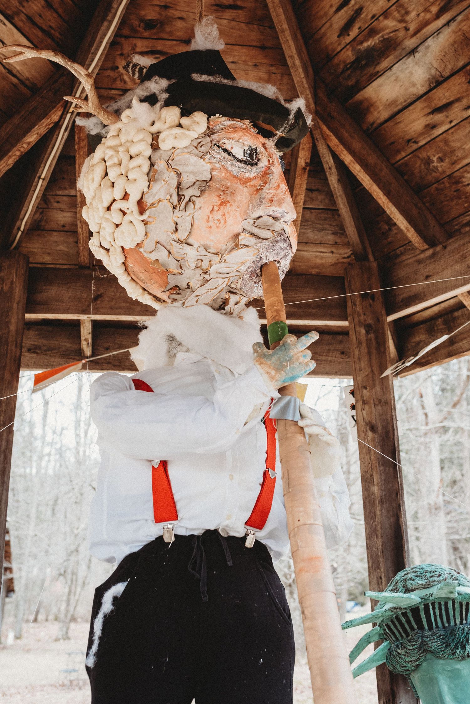 Close-up of handcrafted scarecrow wearing suspenders and white shirt under wooden shelter.