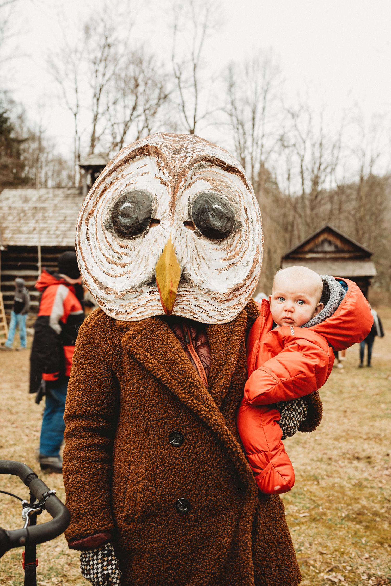Large owl mask costume figure in brown coat holding small child in bright orange winter wear.