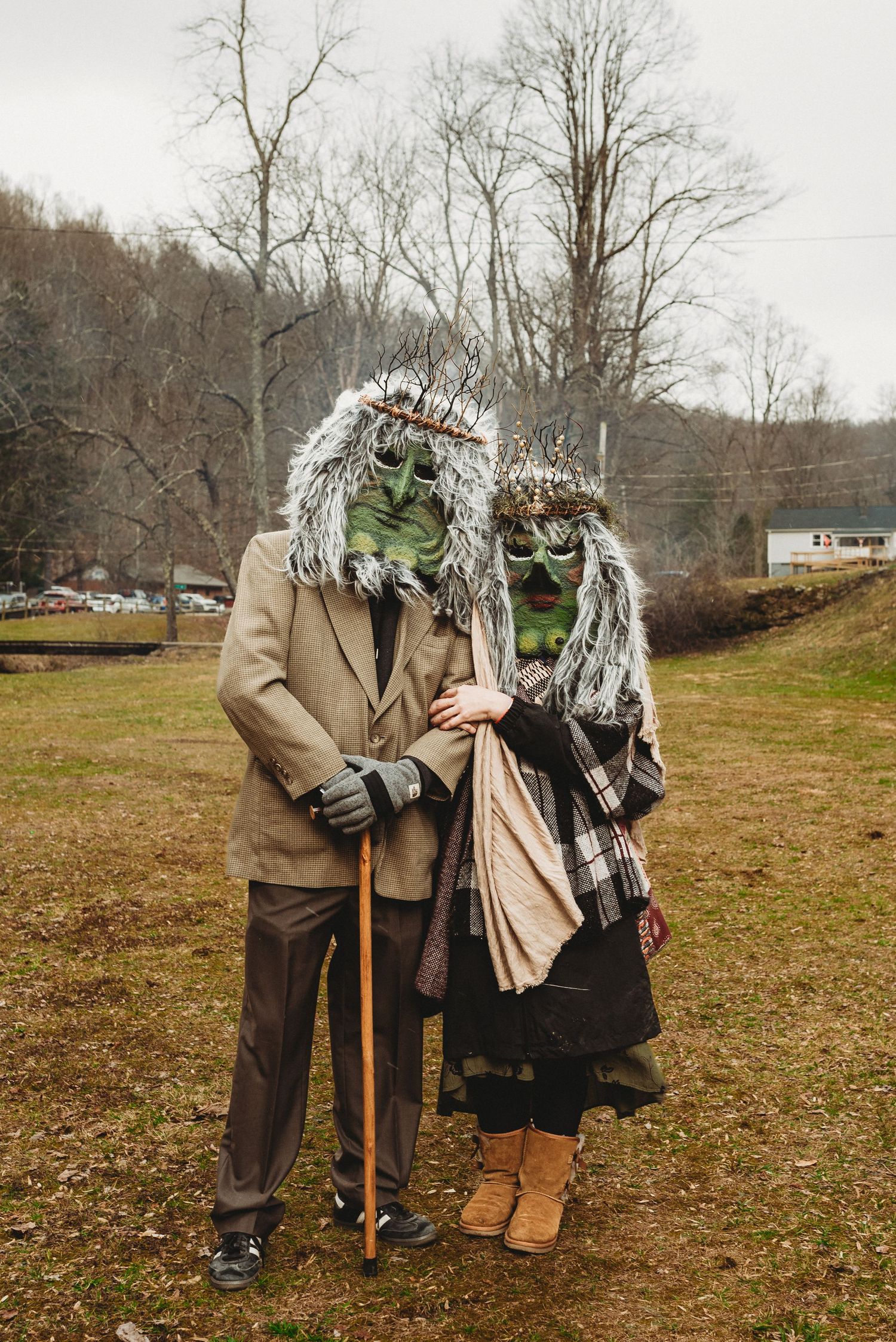 Two figures in vintage clothing and unique bird-like masks standing in a rural field setting.