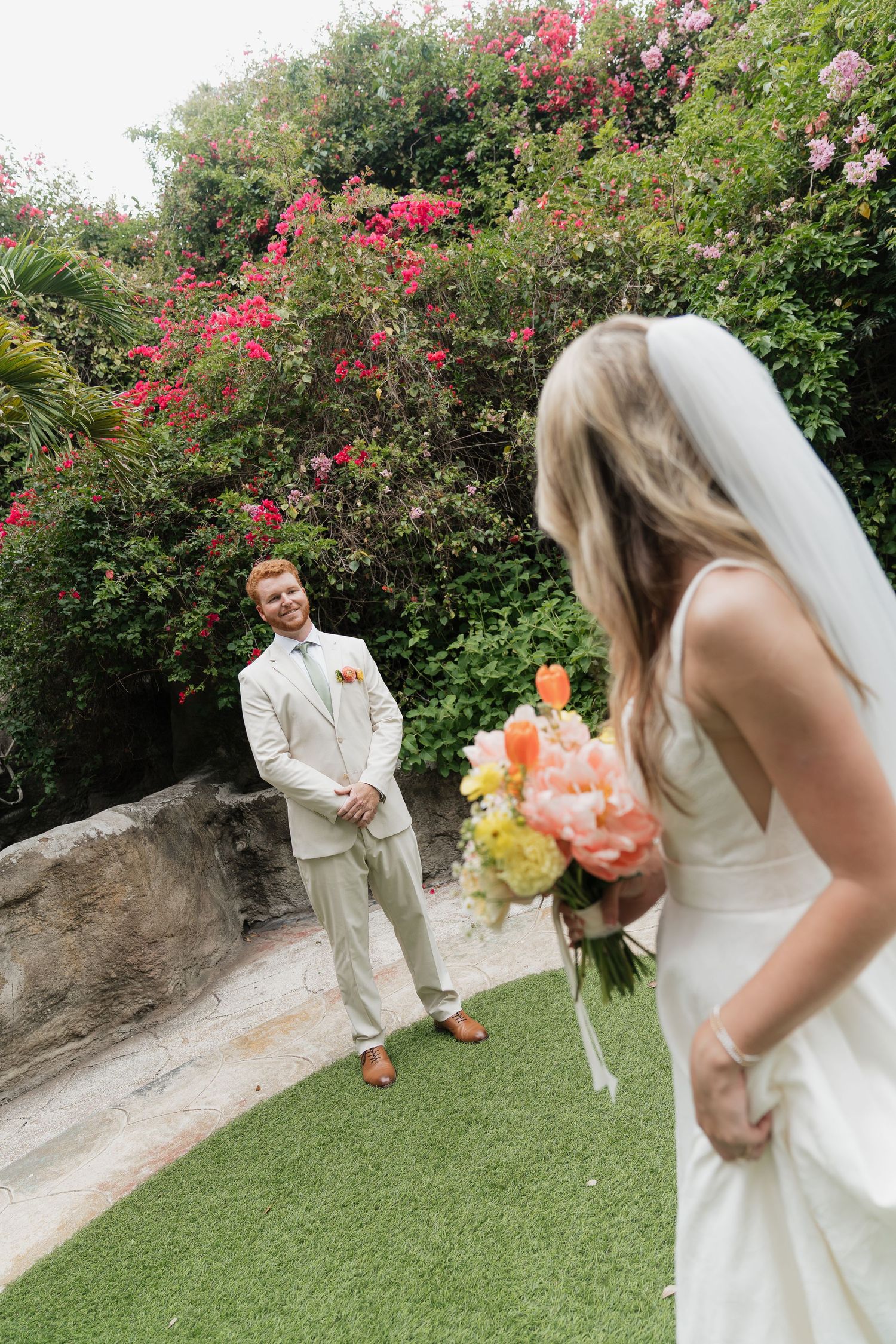 A wedding couple stands in a garden setting with blooming pink flowers and greenery on a sunny day.