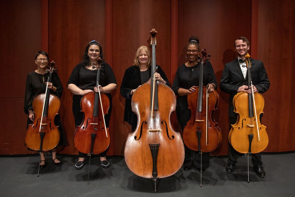 Group portrait of string musicians holding double basses and cellos on stage at a classical concert.